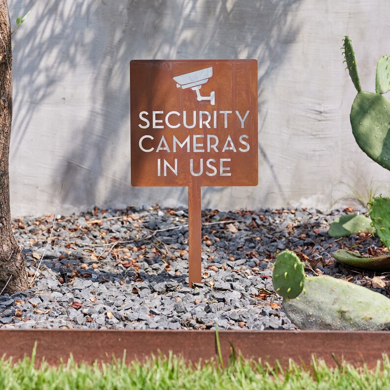 May include: A rusty brown metal sign with white text that reads "SECURITY CAMERAS IN USE". A white silhouette of a security camera is above the text. The sign is mounted on a metal stake and is placed in a bed of gravel.