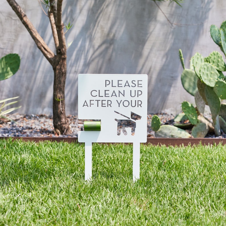 May include: White sign with black text that reads "Please clean up after your" with a silhouette of a dog on a leash. A green plastic bag dispenser is attached to the sign.
