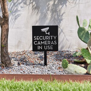May include: Black sign with white text that reads "SECURITY CAMERAS IN USE" with a white silhouette of a security camera above the text. The sign is mounted on a black metal stake and is placed in a bed of gravel.
