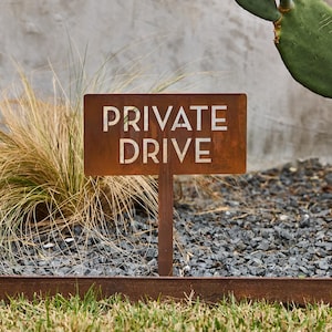 May include: A rectangular, rust-colored metal sign mounted on a vertical stake. The sign features the words "PRIVATE DRIVE" cut out in a bold, sans-serif font, allowing the background to be visible through the lettering.