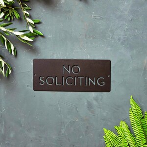 Op de afbeelding: Een zwart metalen bord met de tekst "NO SOLICITING" in witte letters. Het bord is op een grijze achtergrond met groen gebladerte.