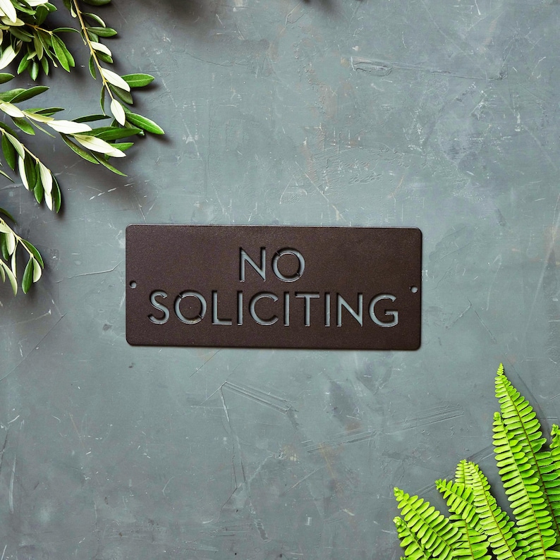 Op de afbeelding: Een zwart metalen bord met de tekst "NO SOLICITING" in witte letters. Het bord is op een grijze achtergrond met groen gebladerte.