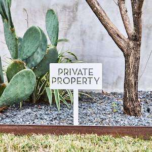 May include: A white rectangular metal sign featuring the cut-out text PRIVATE PROPERTY in a bold, sans-serif font. The sign is mounted on a single vertical stake, positioned upright in a garden bed with gray gravel and green cactus plants.