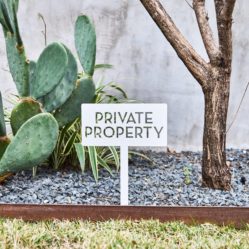 May include: A white rectangular metal sign featuring the cut-out text PRIVATE PROPERTY in a bold, sans-serif font. The sign is mounted on a single vertical stake, positioned upright in a garden bed with gray gravel and green cactus plants.