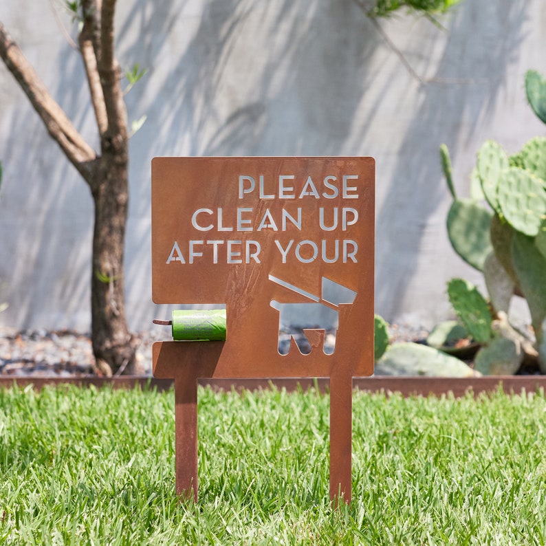 May include: A rusty brown metal sign with the text "PLEASE CLEAN UP AFTER YOUR" and a silhouette of a dog with a leash. A green plastic bag dispenser is attached to the sign.