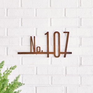 May include: A rusty brown metal house number sign with the number 107. The sign is mounted on a white brick wall. The sign has a horizontal bar with the text "No." and the number "107" in a stylized font.