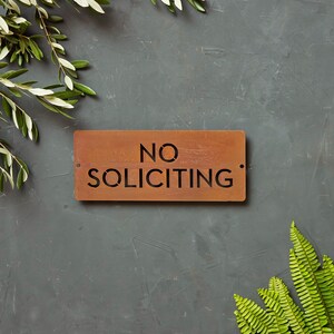 Op de afbeelding: Een roestig metalen bord met de tekst "NO SOLICITING" in zwarte letters. Het bord is op een grijze achtergrond met groen gebladerte aan beide kanten.