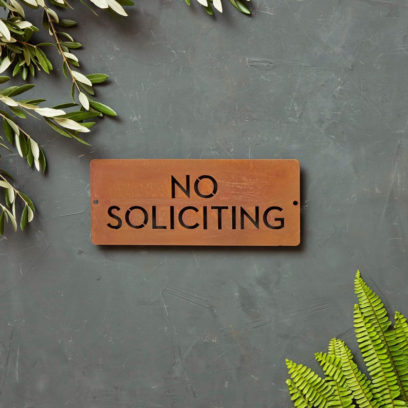 Op de afbeelding: Een roestig metalen bord met de tekst "NO SOLICITING" in zwarte letters. Het bord is op een grijze achtergrond met groen gebladerte aan beide kanten.