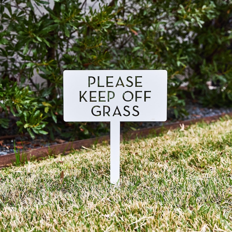 May include: A rectangular white metal sign on a stake featuring the black cut-out text PLEASE KEEP OFF GRASS. The sign is positioned upright in a lawn area, with the text clearly visible in a sans-serif font against the solid white background.