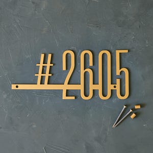 May include: A gold-colored metal house number sign with the number 2605. The sign has a hashtag symbol and includes two screws and two gold-colored screw covers. The sign is on a textured, dark gray background.