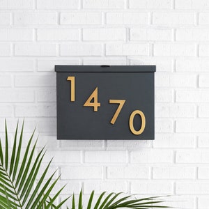 May include: A dark gray mailbox with the numbers "1470" in gold. The mailbox is mounted on a white brick wall. Green palm leaves are visible in the lower left corner.
