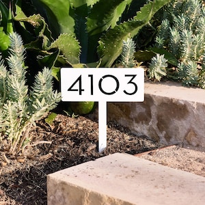 May include: A white rectangular garden stake featuring the black numbers 4103 printed in a clean, modern sans-serif font. The sign is mounted on a vertical post designed to be inserted into the ground, displaying the house number clearly.