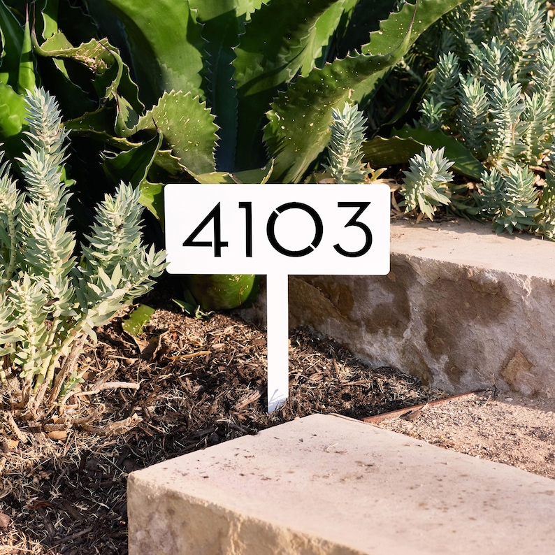 May include: A white rectangular garden stake featuring the black numbers 4103 printed in a clean, modern sans-serif font. The sign is mounted on a vertical post designed to be inserted into the ground, displaying the house number clearly.