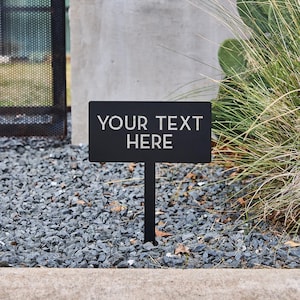 May include: A black rectangular sign with the text "YOUR TEXT HERE" in white lettering. The sign is mounted on a black metal stake and is placed in a bed of gray gravel. Green plants are in the background.