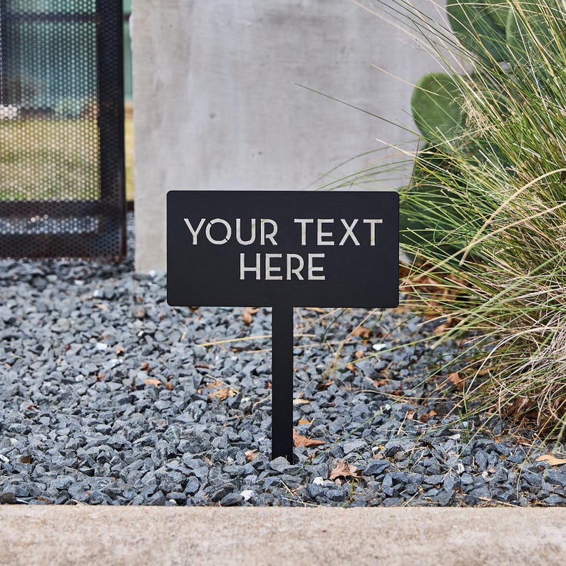 May include: A rectangular black metal garden sign on a thin stake featuring the laser-cut text YOUR TEXT HERE in a clean sans-serif font. The sign is positioned vertically, displaying the cutout lettering clearly against the dark metal surface.