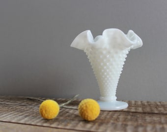 May include: White milk glass bud vase with a ruffled top and a textured, dotted surface. The vase is sitting on a wooden surface with two yellow billy balls.