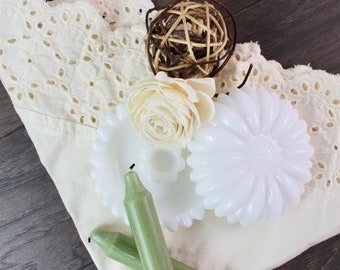May include: White milk glass candle holder with a scalloped edge and a floral design. The holder is on a white crocheted fabric with a brown wooden ball ornament and a white flower.