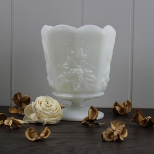 May include: White milk glass vase with a grapevine design. The vase has a pedestal base and is decorated with raised grape clusters and leaves. The vase is surrounded by dried flower pods on a dark wood surface.