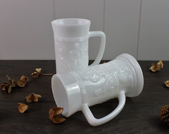 May include: Two white ceramic mugs with raised embossed designs. The mugs have handles and are made of opaque white milk glass.