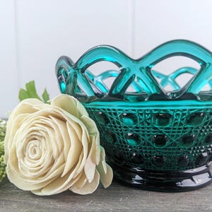 May include: A teal blue glass bowl with a woven design and a textured bottom. The bowl is sitting on a wooden surface next to a white flower.