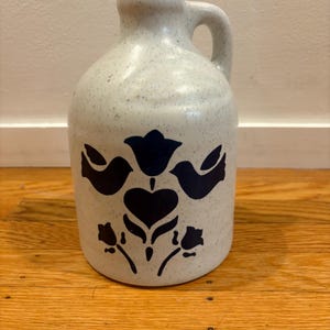 Puede incluir: Jarra de cerámica moteada de color blanquecino con asa. La jarra presenta un diseño floral azul oscuro con dos pájaros y un corazón. La jarra está sobre una superficie de madera.