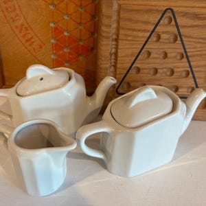 Puede incluir: Un juego de té de cerámica blanca con una tetera, una jarra de crema y un azucarero. La tetera tiene tapa y boquilla. La jarra de crema tiene asa y boquilla. El azucarero tiene tapa.