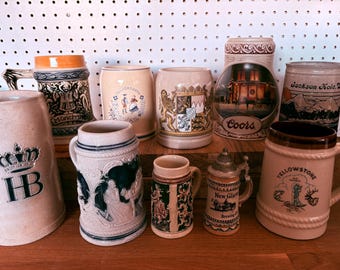 Puede incluir: Una colección de jarras de cerveza vintage de varios tamaños y diseños. Las jarras presentan ilustraciones detalladas, incluyendo escenas, escudos y texto. Los colores incluyen beige, marrón, azul y dorado. Algunas jarras tienen tapas y asas. Las jarras están sobre una superficie de madera.