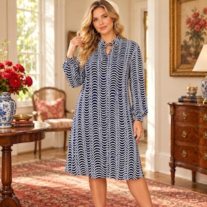 May include: A knee-length dress featuring a navy blue base with a repeating white vertical wavy line pattern. The garment has long sleeves with gathered cuffs, a high neckline with a small keyhole opening, and a tie detail at the collar.