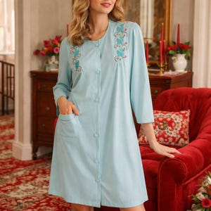 May include: A light blue button-down robe with three-quarter sleeves and two front pockets. The robe features embroidered floral designs in shades of blue and green on the upper chest area. The robe is knee-length.