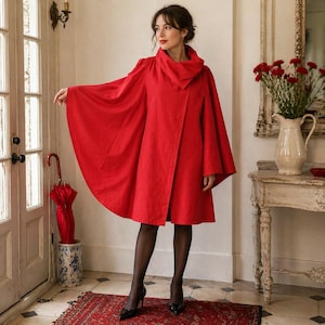 May include: A vibrant red cape coat with a high collar and flowing sleeves. The coat is knee-length and worn with black tights and heels. A red umbrella stands in a decorative holder near a white door.
