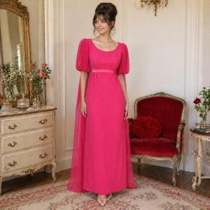 May include: A vibrant fuchsia-colored formal gown with short, puffed sleeves and a flowing cape. The dress features a fitted bodice and a floor-length skirt. A matching ribbon belt cinches the waist. The gown is worn with silver-colored heels.