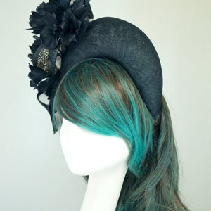 Op de afbeelding: Een brede zwarte hoofdband met een grote zwarte bloem en een zwart en gouden mesh accent. De hoofdband wordt gedragen door een mannequin met turquoise en bruin haar.