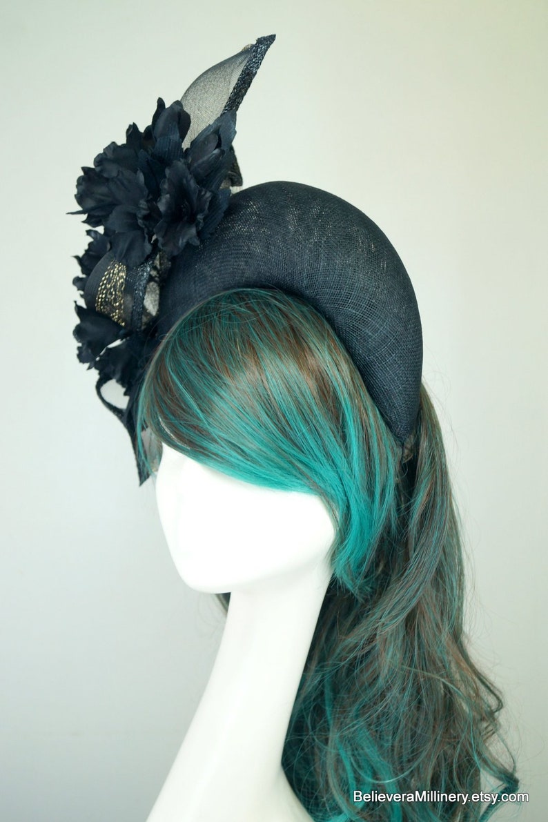 Op de afbeelding: Een brede zwarte hoofdband met een grote zwarte bloem en een zwart en gouden mesh accent. De hoofdband wordt gedragen door een mannequin met turquoise en bruin haar.