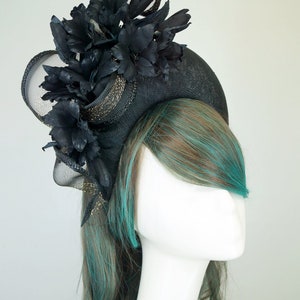 Op de afbeelding: Een zwarte en gouden fascinator met een grote zwarte bloem en gaas. De fascinator wordt gedragen door een mannequin met turquoise en bruin haar.