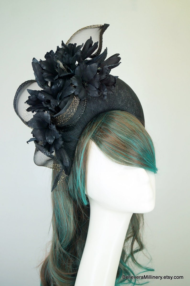 Op de afbeelding: Een zwarte en gouden fascinator met een grote zwarte bloem en gaas. De fascinator wordt gedragen door een mannequin met turquoise en bruin haar.