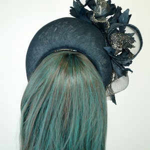 Op de afbeelding: Een zwarte en gouden fascinator met een brede rand en een groot, gekruld verenaccent. De fascinator wordt gedragen door een model met turquoise haar.