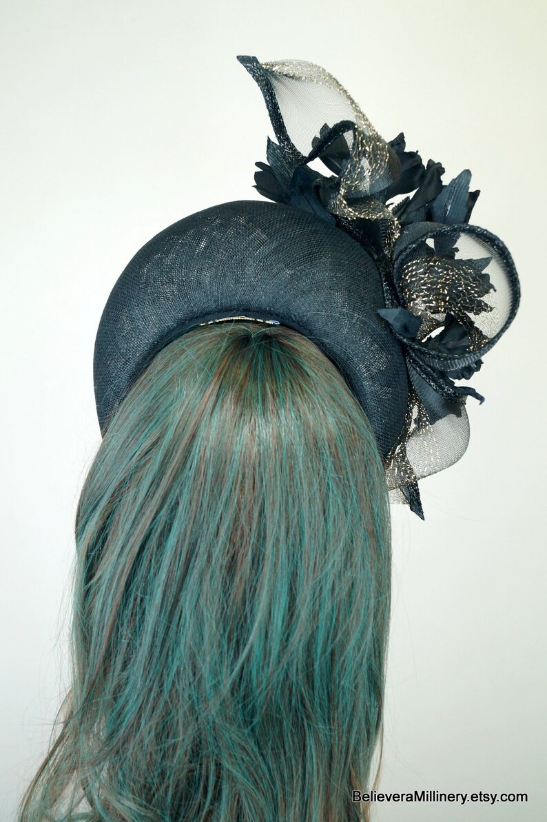 Op de afbeelding: Een zwarte en gouden fascinator met een brede rand en een groot, gekruld verenaccent. De fascinator wordt gedragen door een model met turquoise haar.