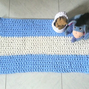 Può includere: Una coperta rettangolare all'uncinetto fatta a mano con strisce alternate azzurre e bianche. Un piccolo cestino all'uncinetto bianco contiene un orsacchiotto marrone e un peluche blu di Eeyore poggia sulla coperta. Un articolo fatto a mano.
