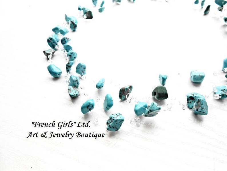 May include: A turquoise gemstone necklace with a clear string. The necklace is on a white surface. The text "French Girls" Ltd. Art & Jewelry Boutique is visible in the image.