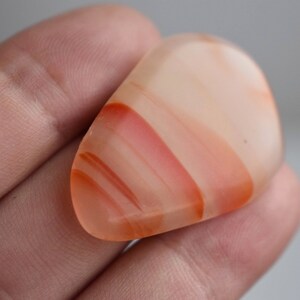 Puede incluir: Una piedra de ágata lisa, translúcida, de color naranja claro con bandas rojas y rosadas. La piedra tiene forma de lágrima y se sostiene en una mano.