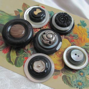 May include: A selection of vintage buttons in diverse designs and colours. The buttons showcase a variety of materials, including black, brown, and white plastic, with decorative metal and mother-of-pearl accents. The buttons are arranged on a floral patterned background.