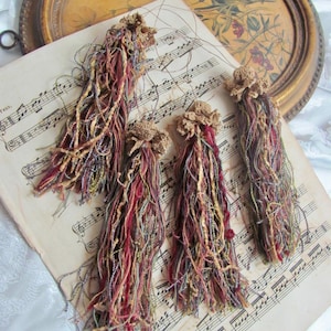 May include: Four handmade tassels with a crochet top and colorful threads in shades of red, brown, and gold. The tassels rest on a vintage sheet of music, with a decorative tray in the background.