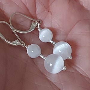 May include: A pair of leverback earrings featuring three white, spherical beads connected by silver-toned metal. The earrings are displayed against a blurred background.