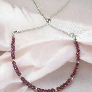 May include: A bracelet with small, round, deep red beads. The bracelet has a silver-coloured chain and a sliding clasp for adjustable sizing. The beads are interspersed with small silver-coloured accents. The bracelet is displayed on a pink fabric.