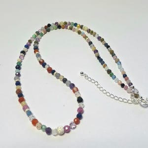 May include: A multi-coloured beaded necklace featuring small, faceted, round gemstones in various hues including blue, red, green, purple, and clear. The necklace includes a silver-toned metal chain extender with a lobster claw clasp closure.