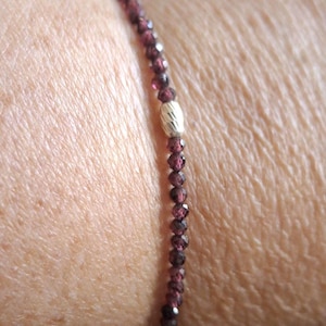 Peut inclure: Un bracelet délicat composé de petites perles rouges foncées et facettées. Une perle argentée texturée est centrée sur le bracelet. Le bracelet est porté sur un poignet, mettant en valeur le design et la couleur du bijou.