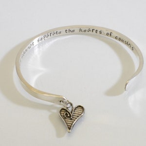 May include: Silver bangle bracelet with a heart charm. The bracelet is engraved with the text "Distance cannot separate the hearts of cousins".