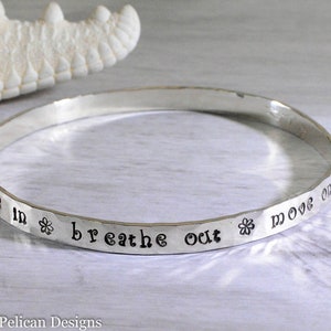 Puede incluir: Pulsera de plata con la inscripción "breathe in breathe out move on" grabada. La pulsera está decorada con pequeños símbolos de flores.