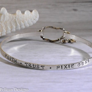 Könnte beinhalten: Ein silbernes Armreif-Armband mit der Gravur "Faith, Trust, Pixie Dust".