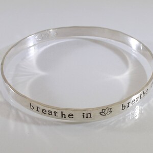 Könnte beinhalten: Silberner Armreif mit der Aufschrift "breathe in breathe out" und einem Lotusblüten-Symbol.